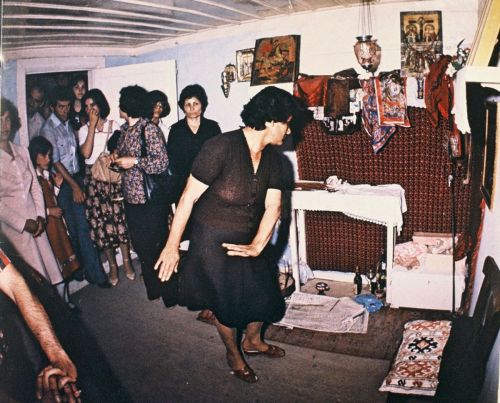 Szenenfoto in Farbe: Eine Gruppe von Menschen in einem Wohnzimmer, einige Tanzen, andere unterhalten sich im Türrahmen.