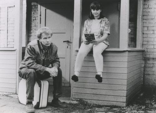 Black and white photo: A man is sitting on a suitcase in front of a house, next to him, a girl is sitting on the rail of the porch and reading a book.