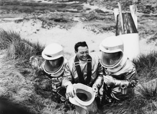 Black and white still: Three men in space suits clumb up a sand dune.
