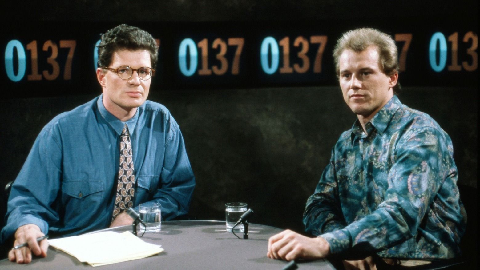 Zwei Männer schauen in die Kamera und sitzen an einem Tisch im Fernsehstudio