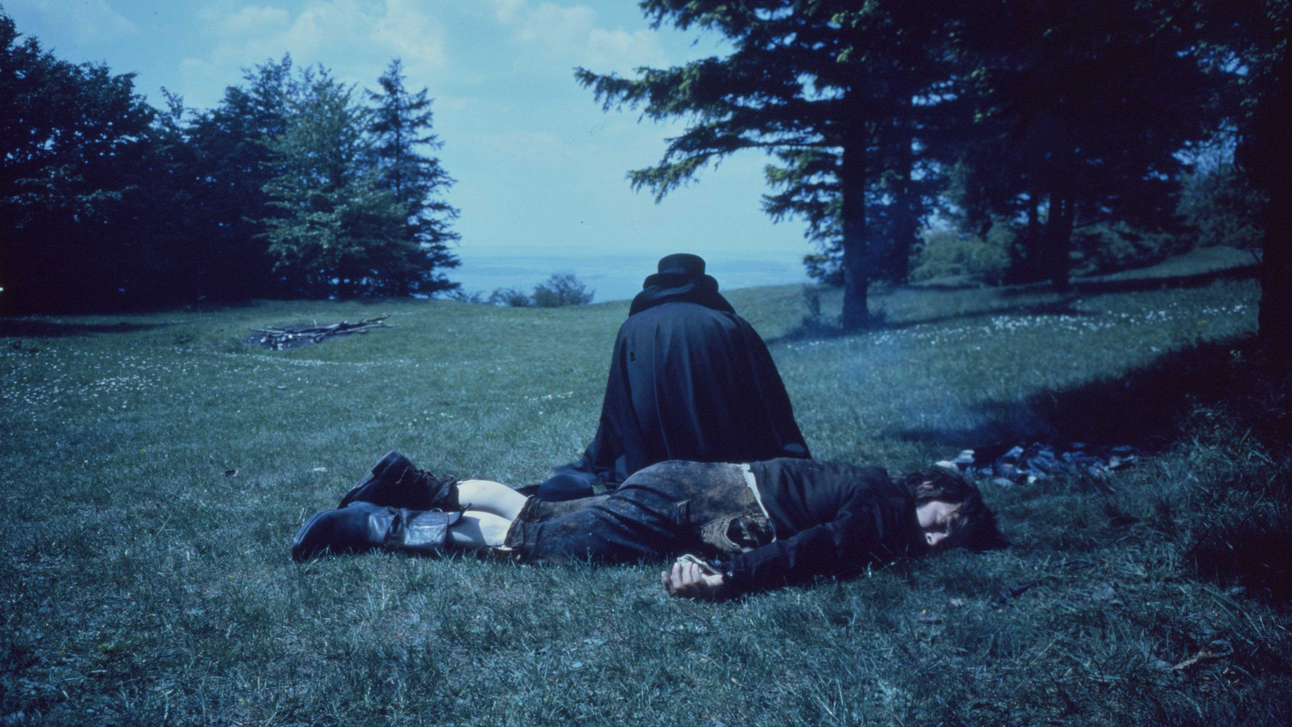 Ein toter Mann liegt auf der Wiese. Hinter ihm hockt ein Mensch in dunkle Kleidung gehüllt, man sieht nur seinen Rücken.