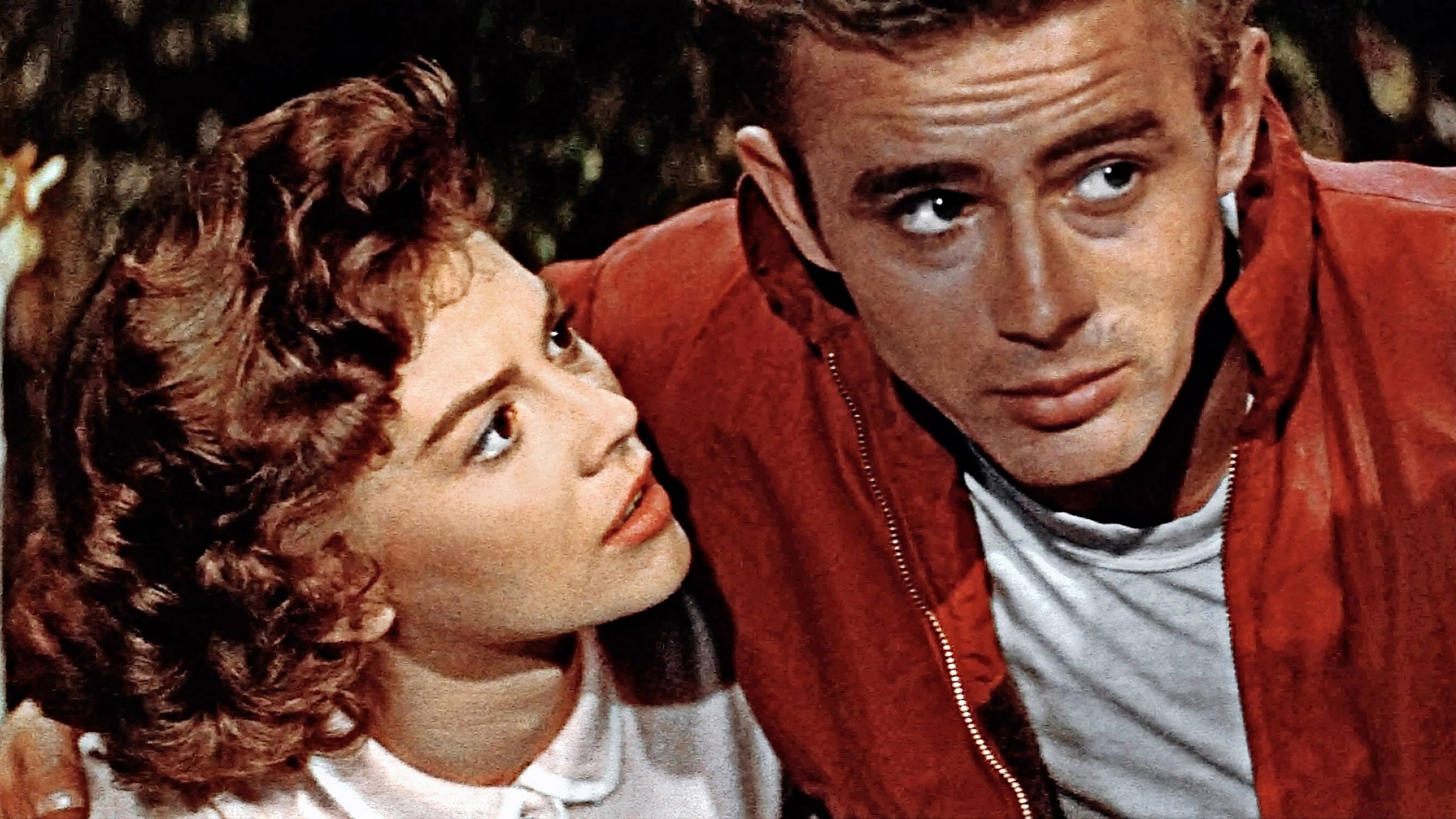 Junger Mann in roter Lederjacke legt den Arm um eine junge Frau, die ihn anblickt.