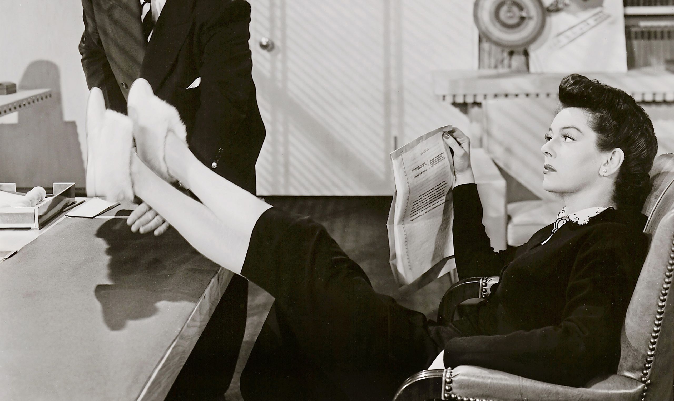 Schwarz-weiß-Szenenfoto: Frau sitzt am Schreibtisch, die Füße mit puscheligen Schuhen lässig auf den Tisch gelegt