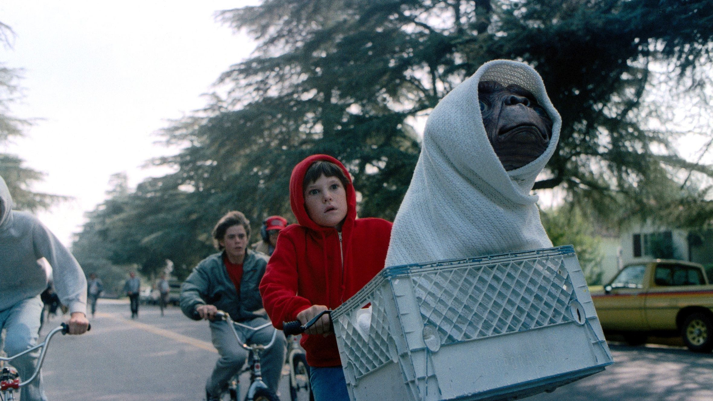 Ein junge in einem roten Kapuzenpullover fährt mit dem Rad und in seinem Korb sitzt ein in eine Decke gewickeltes Alien.