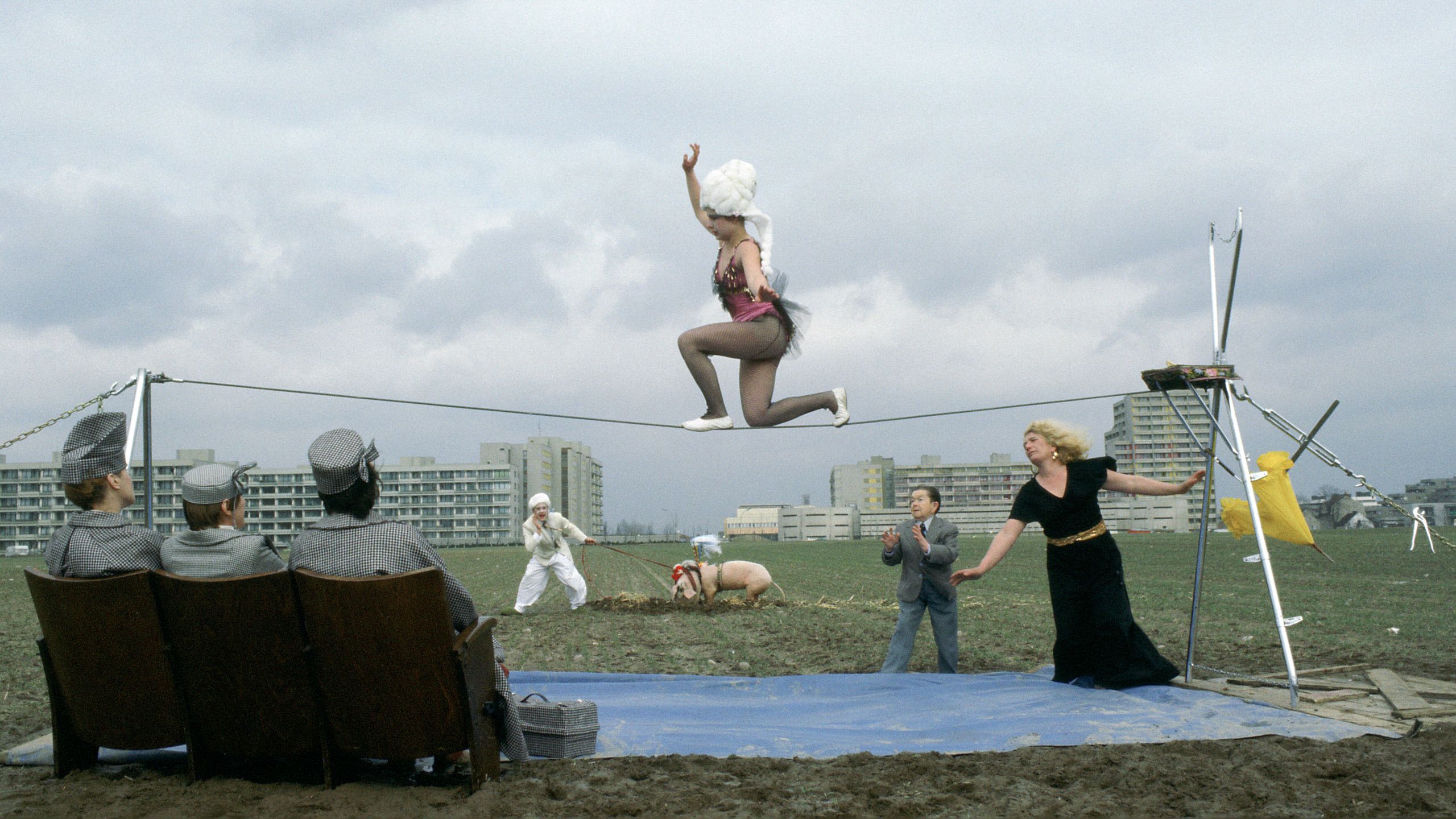Szenenfoto: In einer Berliner Brachlandschaft ist eine Seilkonstruktion aufgebaut, auf der eine kostümierte Seiltänzerin kniet. Im Vordergrund drei Zuschauende in grauer Kleidung auf Sesseln, im Hintergrund große Wohnanlagen