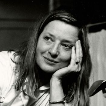 Eine Frau sitzt, den Kopf in die Hand gestützt. Sie lächelt.