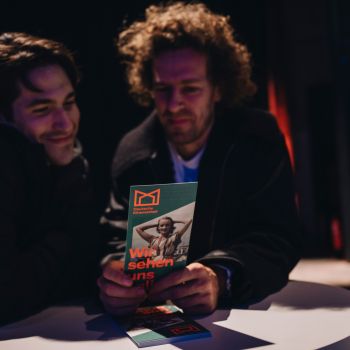 Zwei Männer an einem Stehtisch schauen lächelnd auf einen Flyer.