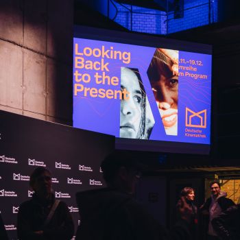 Das Visual zur Filmreihe wird auf einen Screen projiziert, während sich Menschen in der Halle der Kinemathek versammeln.