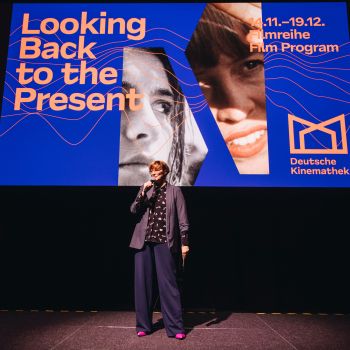 Eine Frau in einem Jacket hält ein Mikrofon und steht vor einer Leinwand auf einer Bühne.