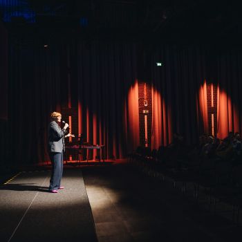 Eine Frau steht mit einem Mikrofon vor einem Publikum in einer halbdunklen Halle.