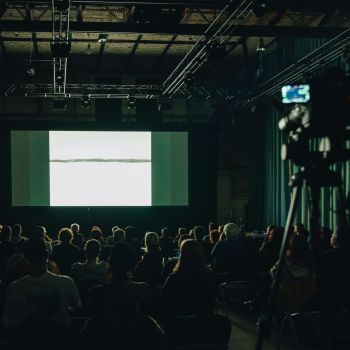 Viele Menschen sitzen in einer Halle vor einer Leinwand, auf der eine weiße Schneelandschaft zu sehen ist.
