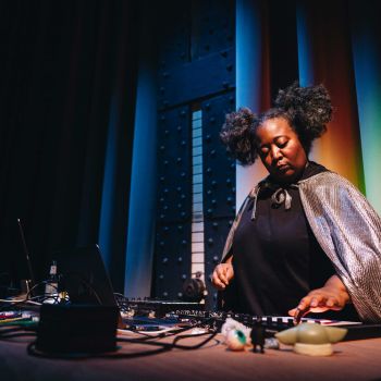 Eine Frau in einem silbernen Cape bedient elektronische Musikmischer, hinter ihr leuchtet buntes Licht.
