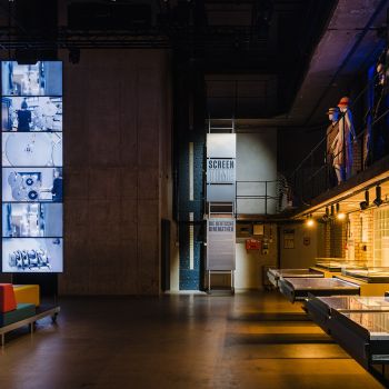 Dunkler Raum mit Sitzgelegenheiten, Vitrinen und Videowand.