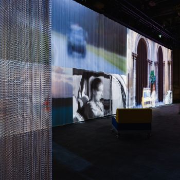 Dunkler Raum mit einem langen gro&szlig;formatigen Band an Filmprojektionen, im Vordergrund ein Kettenvorhang.