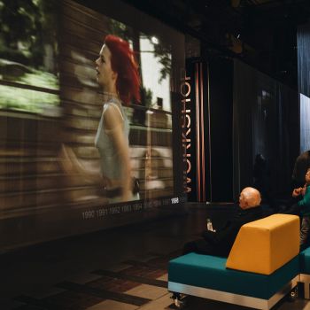 Menschen sitzen in einer Halle auf einer Sofalandschaft und schauen auf eine Leinwand, auf der der Film Lola rennt gezeigt wird.