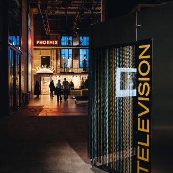 Dunkler Raum mit einer begehbaren Box mit gro&szlig;em Schriftzug TELEVISION im Vordergrund und Menschen im Hintergrund.