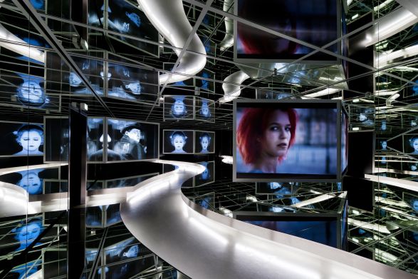 Spiegelnde Ausstellung mit Filmszenen auf Monitoren, leuchtender Weg f&uuml;hrt durch Raum, zentrale Szene zeigt junge Frau mit roten Haaren.