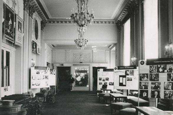 In einem historischen Raum mit Kronleuchter an der Decke stehen zahlreiche Stellw&auml;nde mit Fotografien.