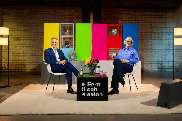 Zwei Personene sitzen auf einem Podium vor einem Hintergrund mit knalligen Farbfl&auml;chen.