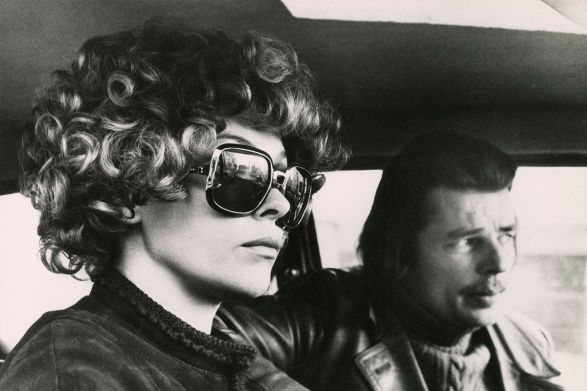 Eine Frau mit gro&szlig;er Sonnenbrille sitzt ernst neben einem Mann mit halblangem Haar und Schnurrbart im Auto.