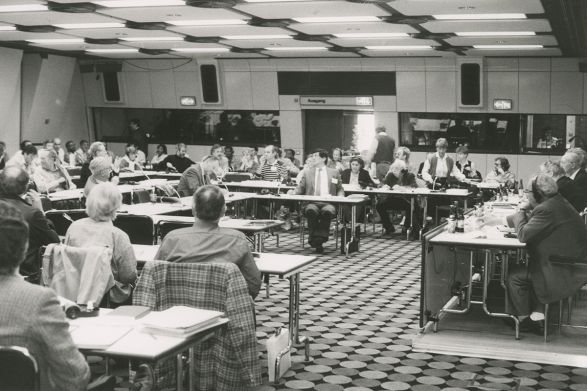 Schwarz-Wei&szlig;-Foto eines Konferenzraums, viele Menschen mit Kopfh&ouml;rern an Tischen, Dolmetscherkabinen im Hintergrund, konzentrierte Stimmung.