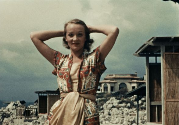 Marlene Dietrich steht in einem buntem Kleid vor Ruinen und H&auml;user. Sie hat die H&auml;nde hinter dem Kopf verschr&auml;nkt.
