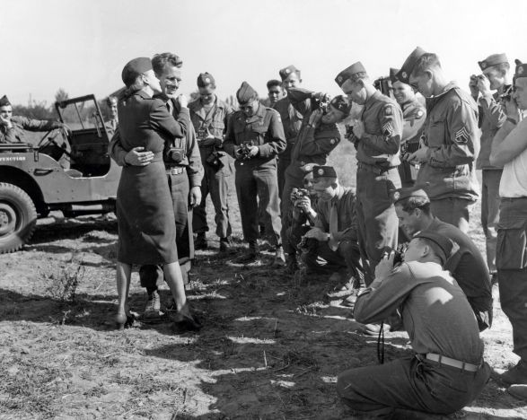 Marlene Dietrich steht im Arm von einem Mann. Viele Soldaten stehen vor den beiden und fotografieren sie. 