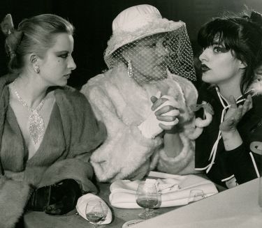 Drei elegant gekleidete Frauen sitzen auf einem Schwarzwei&szlig;foto nebeneinander und trinken Alkohol, eine spricht, zwei h&ouml;ren zu.