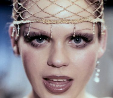Eine stark geschminkte Frau blickt singend in die Kamera. A heavily made-up woman looks singing into the camera.
