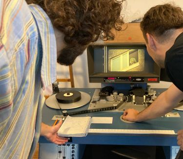 Two men from behind are looking at a screen on which a movie is being played with a projector.