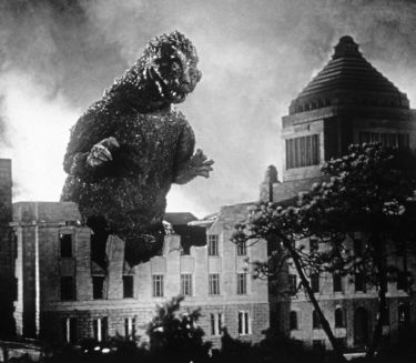 Black and white shot of a huge, lizard-like monster in front of a burning city, smoke and debris in the foreground, skyscrapers visible in the background.