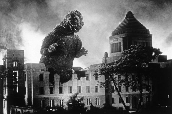 Schwarzweißaufnahme eines riesigen, echsenartigen Monsters vor brennender Stadt, Rauch und Trümmer im Vordergrund, Hochhäuser im Hintergrund sichtbar.