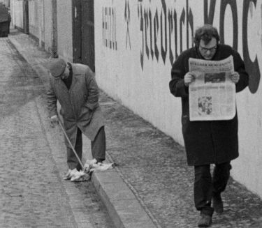 Ein Mann l&auml;uft auf einem B&uuml;rgerstei und liest die Zeitung, w&auml;hrend ein anderer Mann in der N&auml;he mit seinem Spazierstock in einer fallengelassenen Zeitung stochert.