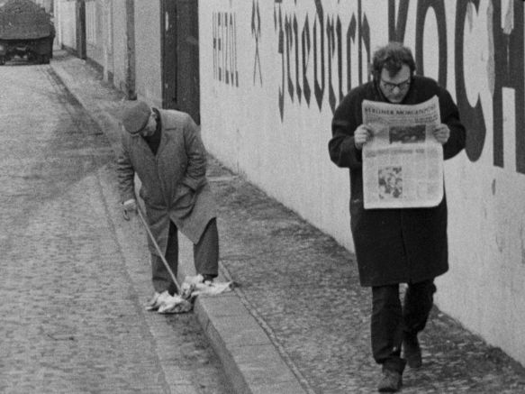 Ein Mann läuft auf einem Bürgerstei und liest die Zeitung, während ein anderer Mann in der Nähe mit seinem Spazierstock in einer fallengelassenen Zeitung stochert.