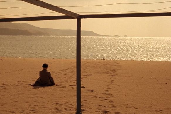 Eine ältere Frau mit Hut sitzt am Strand, Rücken zur Kamera, und schaut aufs Meer.