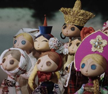 Szenenfoto aus einem Puppentrickfilm: Eine Gruppe von jungen Frauen in folkloristischer Kleidung blickt nach links auf ein Ereignis au&szlig;erhalb des Bildraums.
