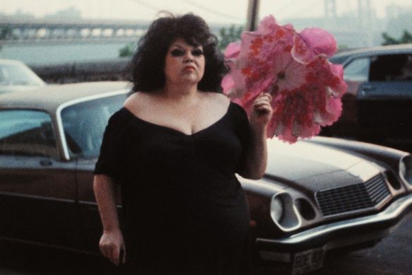 Eine stark geschminkte Frau im schwarzen Kleid steht mit pinkem Regenschirm vor einem Cadillac.