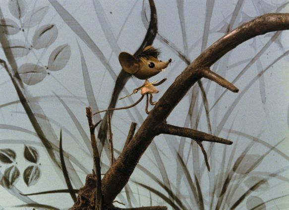 Ein Maus aus Holz läuft auf einem Zweig, im Hintergrund Gräser und Blätter.