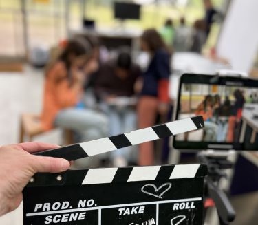 Im Vordergrund eine Hand, die eine Filmklappe h&auml;lt, im Hintergrund (unscharf) ein Tablet und eine Gruppe von Kindern.
