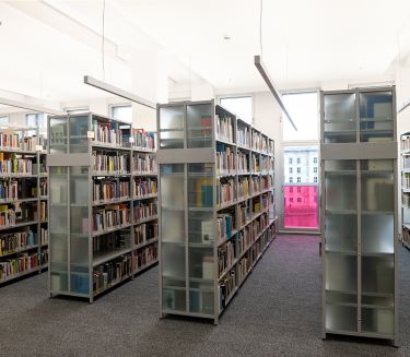 Several shelves with books are located in the library in front of a large window wall.