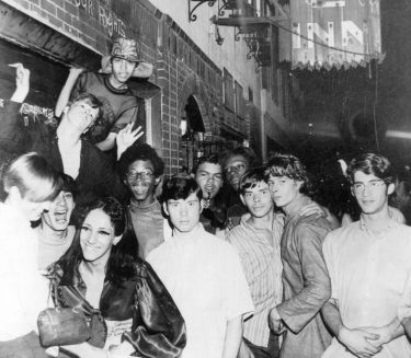 Eine Gruppe junger Menschen vor einem Geb&auml;ude mit einem beleuchteten Schild.