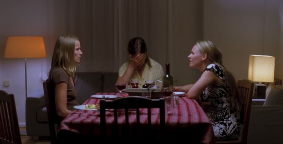 Drei Frauen sitzen an einem Tisch. Die mittlere hält sich die Hände vors Gesicht, während die anderen beiden sich wütend ins Gesicht schauen. Die Stimmung scheint angespannt. 