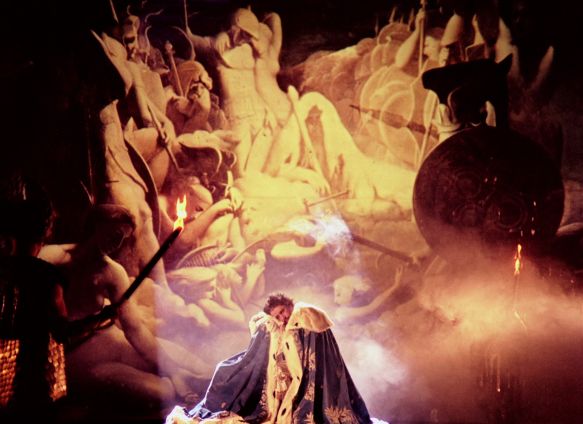 Ein Mann in k&ouml;niglicher Kleidung steht in einem Mantel vor einer Wand mit Soldatenfiguren und nackten Frauen.