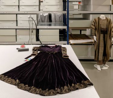 Ein dunkles Samtkleid mit Goldstickereien liegt ausgebreitet auf einem Tisch. Im Hintergrund Regale mit Archivk&auml;sten.