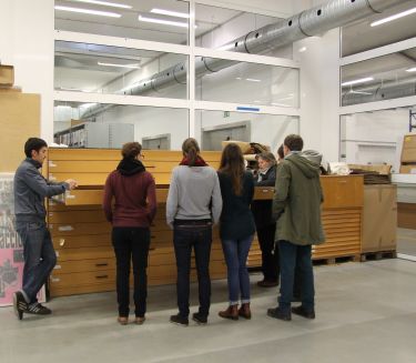 Rechts im Bild erkl&auml;rt eine Archivmitarbeitende einer Gruppe von Besuchenden Plakate aus einem ge&ouml;ffneten Planschrank.