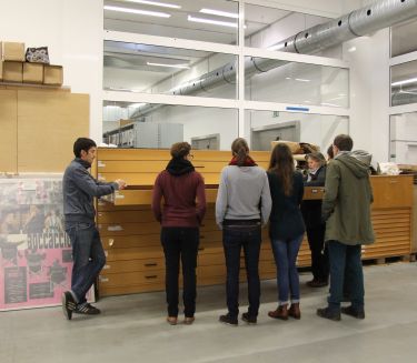 On the right, an archive employee explains posters from an open plan cabinet to a group of visitors.