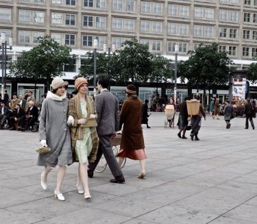 People in 1920s attire stroll across a lively square with modern buildings, trees, and shopfronts in the background.