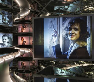 Ausstellungsraum mit beleuchteter Laufbahn und Filmprojektionen, Frauenportr&auml;ts in Farbe und Schwarz-Wei&szlig;, cineastische Atmosph&auml;re mit Spiegeln.