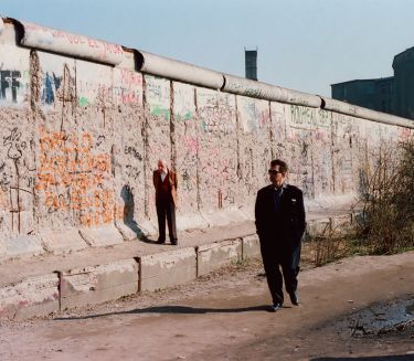 Mann im schwarzen Anzug und Sonnenbrille geht entlang der graffitibedeckten Berliner Mauer, w&auml;hrend ein anderer in einem leuchtend Anzug im Hintergrund steht.