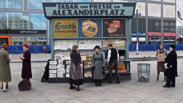 Schwarz-Wei&szlig;-Foto eines Kiosks &raquo;Tabak und Presse&laquo; am Alexanderplatz, mit drei Personen, Zeitungen, Zigaretten und Bus im Hintergrund.
